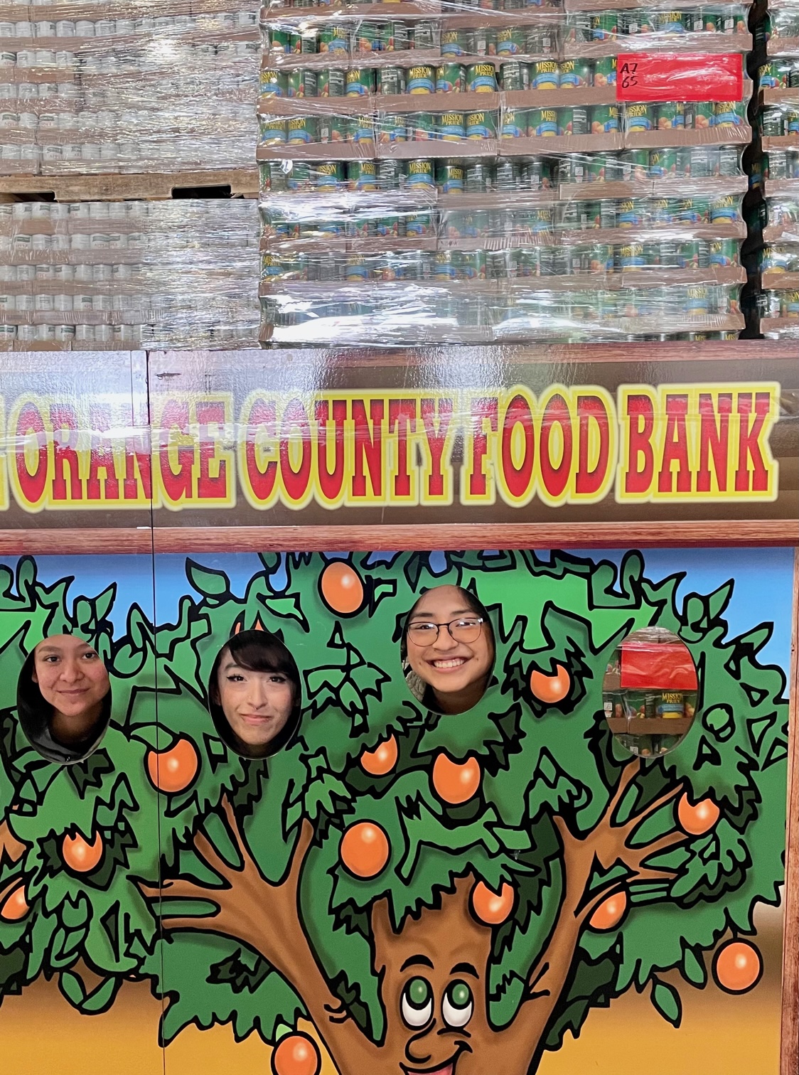 Three SAC ULink students posing for a Orange County Food Bank picture cut-out of a tree with oranges.
